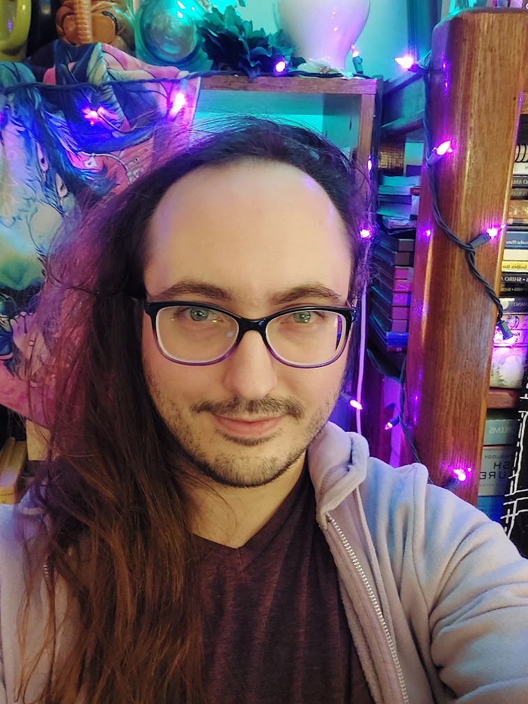 Headshot of Lavra Tamutus backed by a full bookshelf, purple string lights, and a hanging pink bandana adorned by a fluorescent green goat